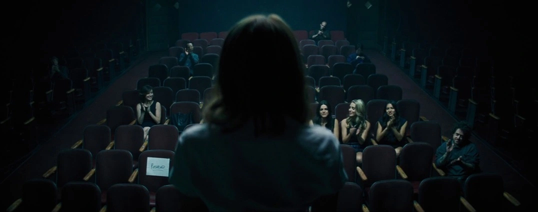A woman sitting in a dim auditorium surrounded by seated audience members. A special seat is empty. From the movie 'La La Land' by Damien Chazelle.